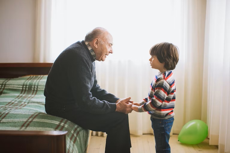 grandfather sitting on the bed talking to standing grandson while holding hands