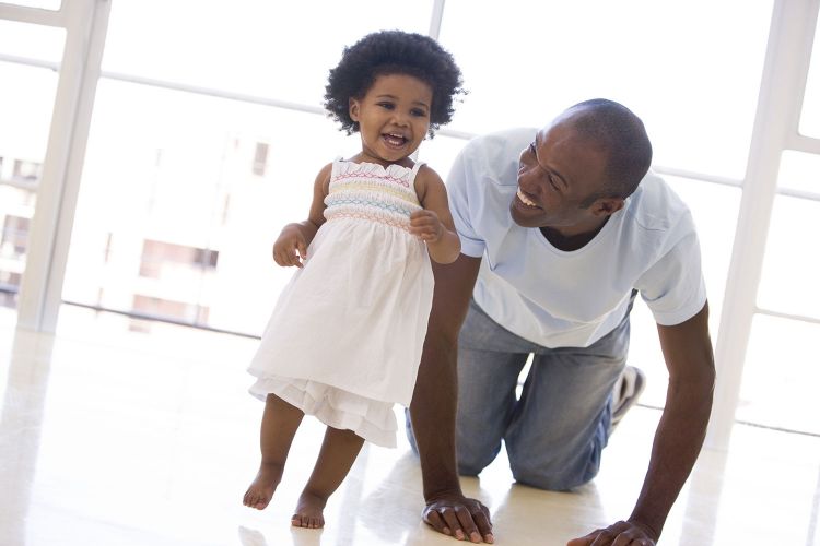 Father is playing with their young toddler daughter, crawling behind her as she runs and laughs.