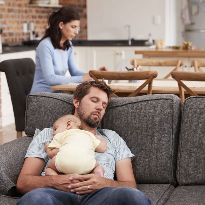 father sleeping on the couch with baby sleeping in his arms and mother working at the table.